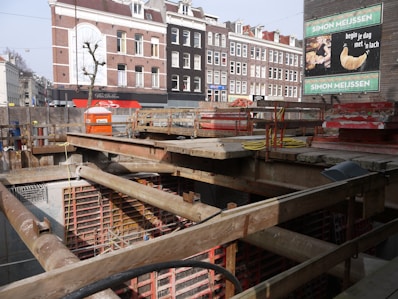 A construction site with a pit and wooden beams across the top. In the background, there are several multi-story brick buildings with a mix of white and dark facades. A billboard advertises a bakery with an image of a croissant.