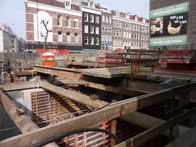 A construction site with a pit and wooden beams across the top. In the background, there are several multi-story brick buildings with a mix of white and dark facades. A billboard advertises a bakery with an image of a croissant.