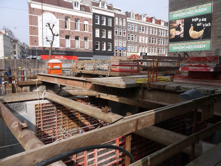 A construction site with a pit and wooden beams across the top. In the background, there are several multi-story brick buildings with a mix of white and dark facades. A billboard advertises a bakery with an image of a croissant.