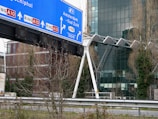 A large blue road sign is displayed, directing towards various routes including Amsterdam and Schiphol. There is modern architecture in the background with glass-paneled buildings and bare trees in the foreground.