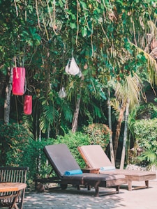Two lounge chairs with blue towels rest on a patio surrounded by lush greenery, including palm trees and shrubs. Several hanging decorative lamps and lanterns add to the ambiance, creating a serene and tropical environment.