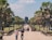 A large stone pathway leads towards an ancient stone temple structure surrounded by lush green palm trees. Several people are walking in both directions, some in casual attire, observing the surroundings. The sky is clear and blue, contributing to the warm and inviting atmosphere.