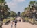 A large stone pathway leads towards an ancient stone temple structure surrounded by lush green palm trees. Several people are walking in both directions, some in casual attire, observing the surroundings. The sky is clear and blue, contributing to the warm and inviting atmosphere.