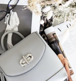A stylish setup includes a light gray handbag with a silver clasp resting on open fashion magazines. Nearby, a pair of black-rimmed glasses adds to the sophisticated look. The scene is complemented by a bouquet of dried flowers and a leopard print fabric, enhancing the fashionable aesthetic.