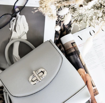 A stylish setup includes a light gray handbag with a silver clasp resting on open fashion magazines. Nearby, a pair of black-rimmed glasses adds to the sophisticated look. The scene is complemented by a bouquet of dried flowers and a leopard print fabric, enhancing the fashionable aesthetic.