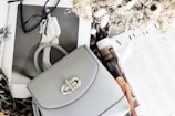 A stylish setup includes a light gray handbag with a silver clasp resting on open fashion magazines. Nearby, a pair of black-rimmed glasses adds to the sophisticated look. The scene is complemented by a bouquet of dried flowers and a leopard print fabric, enhancing the fashionable aesthetic.