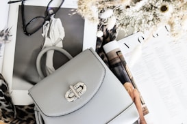 A stylish setup includes a light gray handbag with a silver clasp resting on open fashion magazines. Nearby, a pair of black-rimmed glasses adds to the sophisticated look. The scene is complemented by a bouquet of dried flowers and a leopard print fabric, enhancing the fashionable aesthetic.
