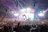 A vibrant concert scene with a large crowd of people raising their hands and taking photos. Colorful lights and confetti enhance the lively atmosphere, while large screens display visuals in the background.
