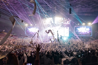 A vibrant concert scene with a large crowd of people raising their hands and taking photos. Colorful lights and confetti enhance the lively atmosphere, while large screens display visuals in the background.