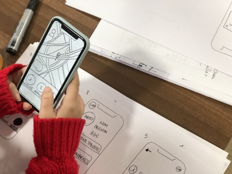 A person wearing red knitted fingerless gloves is holding a smartphone displaying a simple wireframe interface with drawn elements. The phone is placed over sheets of paper containing sketches of app interfaces and handwritten notes. A black marker is visible on the table beside the sketches.