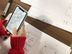A person wearing red knitted fingerless gloves is holding a smartphone displaying a simple wireframe interface with drawn elements. The phone is placed over sheets of paper containing sketches of app interfaces and handwritten notes. A black marker is visible on the table beside the sketches.