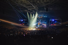 A large indoor concert hall filled with a crowd of people gathered around a brightly lit stage. Multiple beams of colorful stage lights and spotlights illuminate the scene, creating a dynamic and vibrant atmosphere. Large screens on either side of the stage display visuals for the audience.