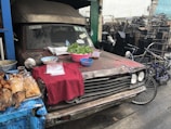 An old, rusty vehicle is parked on the side of the street, with its hood being used as a makeshift table. Various food items, including grilled fish, snacks, and vegetables, are displayed on a red cloth. A bicycle is parked nearby, and the background shows stacks of mechanical parts.