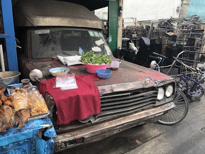 An old, rusty vehicle is parked on the side of the street, with its hood being used as a makeshift table. Various food items, including grilled fish, snacks, and vegetables, are displayed on a red cloth. A bicycle is parked nearby, and the background shows stacks of mechanical parts.