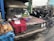 An old, rusty vehicle is parked on the side of the street, with its hood being used as a makeshift table. Various food items, including grilled fish, snacks, and vegetables, are displayed on a red cloth. A bicycle is parked nearby, and the background shows stacks of mechanical parts.