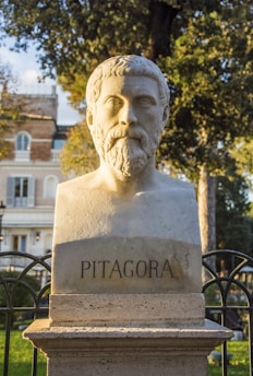 A marble bust of a bearded man with curly hair is set against the backdrop of a garden with trees and a building in the distance. The name 'Pitagora' is inscribed on the pedestal of the bust. The lighting suggests it is either early morning or late afternoon, casting a warm glow on the statue.