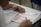 A person is seated at a desk, filling out a standardized test or exam form. The paper has multiple-choice questions and sections for responses. The individual appears focused, using a pen to mark answers.