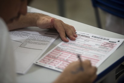 A person is seated at a desk, filling out a standardized test or exam form. The paper has multiple-choice questions and sections for responses. The individual appears focused, using a pen to mark answers.