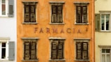 A facade of an aged building with an orange exterior and several wooden shuttered windows, each displaying signs of weathering. The word 'FARMACIA' is prominently displayed in the center, indicating it is or was a pharmacy. Adjacent buildings with different colored exteriors and shutters are also partially visible.