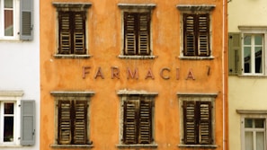 A facade of an aged building with an orange exterior and several wooden shuttered windows, each displaying signs of weathering. The word 'FARMACIA' is prominently displayed in the center, indicating it is or was a pharmacy. Adjacent buildings with different colored exteriors and shutters are also partially visible.