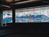 An airplane is docked at an airport gate with people boarding through a jet bridge. Large windows frame the scene, showcasing a backdrop of majestic mountains under a partly cloudy sky.