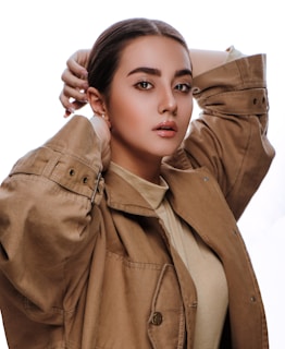 A portrait of a person with dark hair neatly tied back, wearing a light brown jacket with metallic buttons. The pose includes one hand positioned behind the head, conveying a confident and poised demeanor. The facial expression is calm and self-assured.