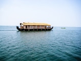 A traditional houseboat with a thatched roof floats on a vast expanse of calm water under a clear blue sky. The boat is elaborately designed with windows and intricate patterns along the sides. There are a few people visible inside, enjoying the serene surroundings.