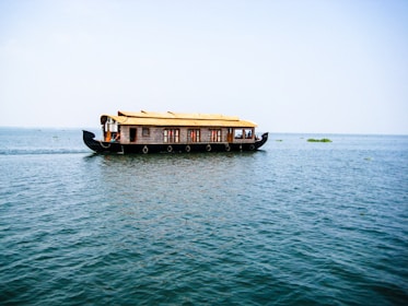 A traditional houseboat with a thatched roof floats on a vast expanse of calm water under a clear blue sky. The boat is elaborately designed with windows and intricate patterns along the sides. There are a few people visible inside, enjoying the serene surroundings.