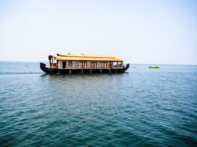 A traditional houseboat with a thatched roof floats on a vast expanse of calm water under a clear blue sky. The boat is elaborately designed with windows and intricate patterns along the sides. There are a few people visible inside, enjoying the serene surroundings.