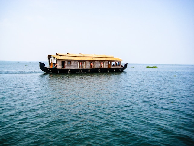 A traditional houseboat with a thatched roof floats on a vast expanse of calm water under a clear blue sky. The boat is elaborately designed with windows and intricate patterns along the sides. There are a few people visible inside, enjoying the serene surroundings.