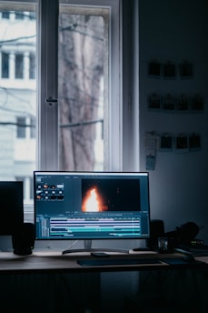 A computer monitor displaying video editing software with a burning flame on the screen. The setup is in a dimly lit room with a wooden desk, keyboard, and window in the background. There are photographs hanging on the wall.