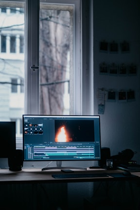 A computer monitor displaying video editing software with a burning flame on the screen. The setup is in a dimly lit room with a wooden desk, keyboard, and window in the background. There are photographs hanging on the wall.