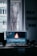 A computer monitor displaying video editing software with a burning flame on the screen. The setup is in a dimly lit room with a wooden desk, keyboard, and window in the background. There are photographs hanging on the wall.