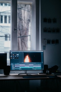 A computer monitor displaying video editing software with a burning flame on the screen. The setup is in a dimly lit room with a wooden desk, keyboard, and window in the background. There are photographs hanging on the wall.
