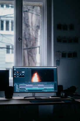 A computer monitor displaying video editing software with a burning flame on the screen. The setup is in a dimly lit room with a wooden desk, keyboard, and window in the background. There are photographs hanging on the wall.