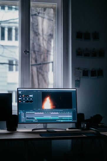 A computer monitor displaying video editing software with a burning flame on the screen. The setup is in a dimly lit room with a wooden desk, keyboard, and window in the background. There are photographs hanging on the wall.