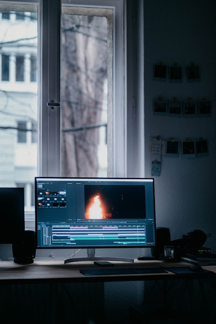 A computer monitor displaying video editing software with a burning flame on the screen. The setup is in a dimly lit room with a wooden desk, keyboard, and window in the background. There are photographs hanging on the wall.