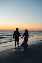 photography of man and woman holding hands each other while walking beside seashore