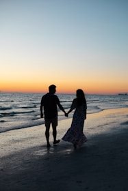 photography of man and woman holding hands each other while walking beside seashore