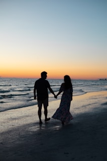 photography of man and woman holding hands each other while walking beside seashore