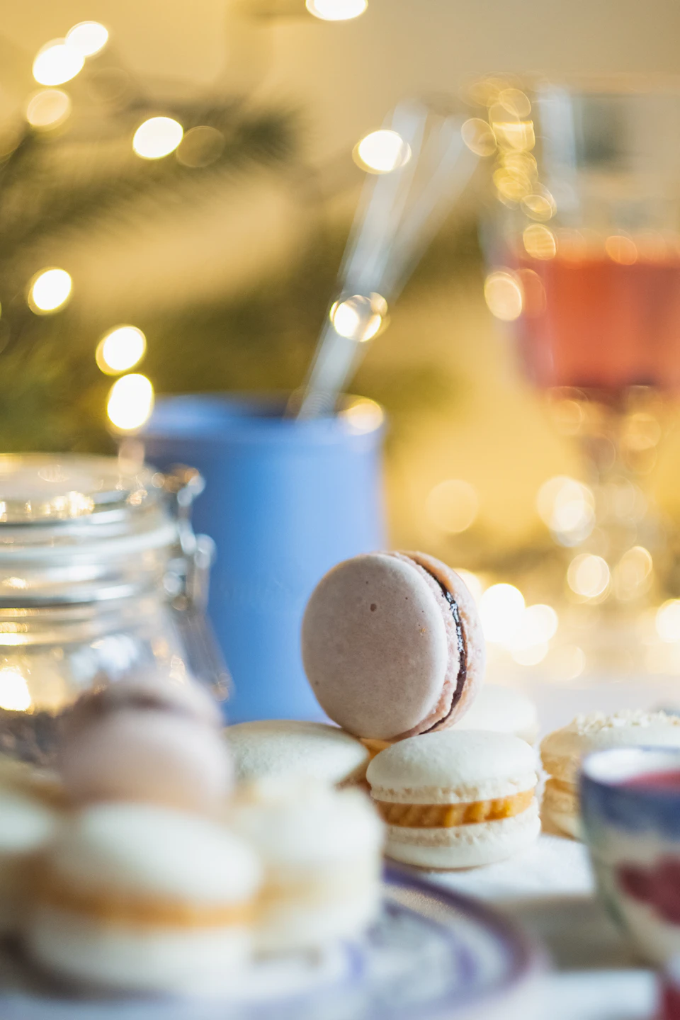 The image shows a table set with various desserts and drinks, including a plate of macarons, a blue mug filled with coffee or tea, and a glass of wine. The macarons are arranged on a white plate, which is placed on a white tablecloth. In the background, there is a blurred Christmas tree, adding to the festive atmosphere.