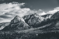 A black and white photograph of a rugged mountain range with sharp peaks and rocky formations. The sky above is partially cloudy, casting varying shadows on the mountains. Dense forests cover the lower slopes, creating a textured landscape.