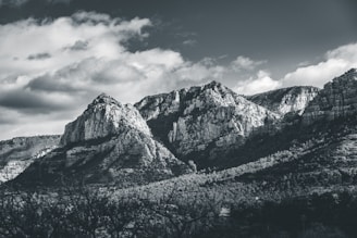 A black and white photograph of a rugged mountain range with sharp peaks and rocky formations. The sky above is partially cloudy, casting varying shadows on the mountains. Dense forests cover the lower slopes, creating a textured landscape.