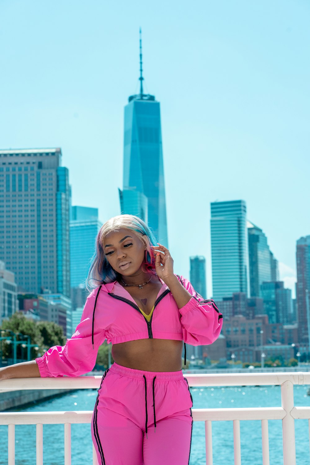 woman wearing pink zip-up crop top and pink bottoms standing and leaning on white railings viewing body of water and high-rise buildings