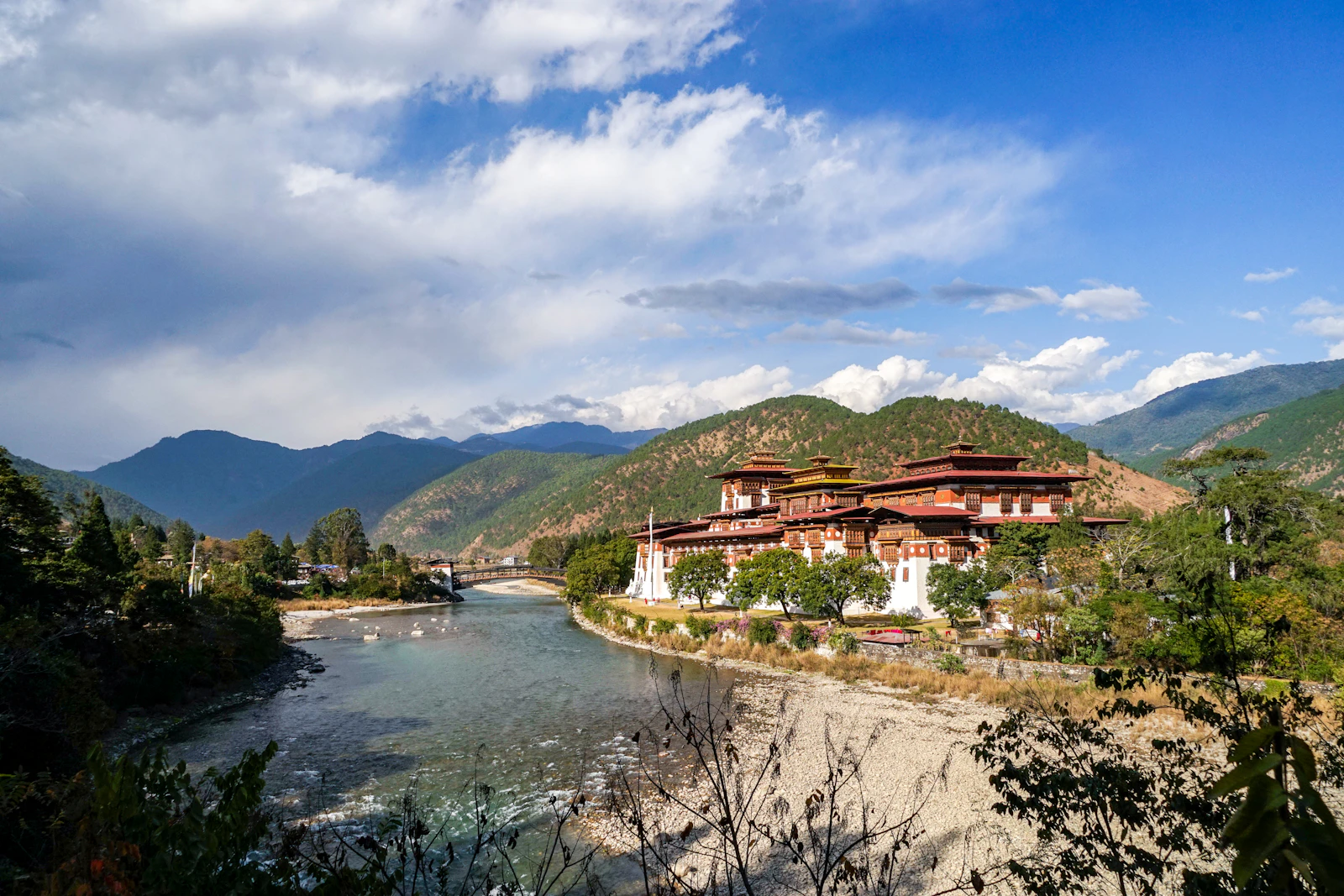 Punakha Dzong beside a clear green river, with a covered bridge and forested hills under cloud
