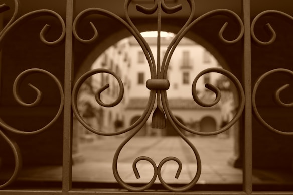 A wrought iron gate with intricate swirling patterns is in the foreground, partially obscuring the view of a courtyard with arches and the facade of a building in the background. The entire scene is colored in sepia tones, giving it an antique appearance.