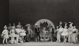 A black and white photograph featuring a group of performers in various costumes. There are people dressed as animals and others in theatrical outfits, such as tutus and top hats. The setting appears to be a stage with a large backdrop in the center, possibly depicting a leaf or abstract design. The group is arranged in a semicircle, showcasing a range of facial expressions and poses.