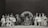 A black and white photograph featuring a group of performers in various costumes. There are people dressed as animals and others in theatrical outfits, such as tutus and top hats. The setting appears to be a stage with a large backdrop in the center, possibly depicting a leaf or abstract design. The group is arranged in a semicircle, showcasing a range of facial expressions and poses.