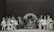 A black and white photograph featuring a group of performers in various costumes. There are people dressed as animals and others in theatrical outfits, such as tutus and top hats. The setting appears to be a stage with a large backdrop in the center, possibly depicting a leaf or abstract design. The group is arranged in a semicircle, showcasing a range of facial expressions and poses.