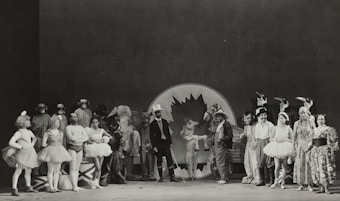 A black and white photograph featuring a group of performers in various costumes. There are people dressed as animals and others in theatrical outfits, such as tutus and top hats. The setting appears to be a stage with a large backdrop in the center, possibly depicting a leaf or abstract design. The group is arranged in a semicircle, showcasing a range of facial expressions and poses.